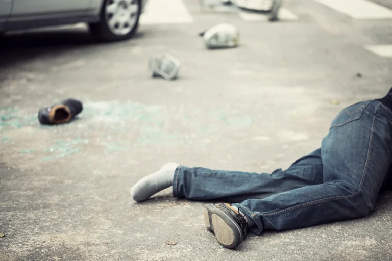 A person lies on the ground next to scattered debris and broken glass on a street, suggesting a serious pedestrian accident with a car and more debris in the background.
