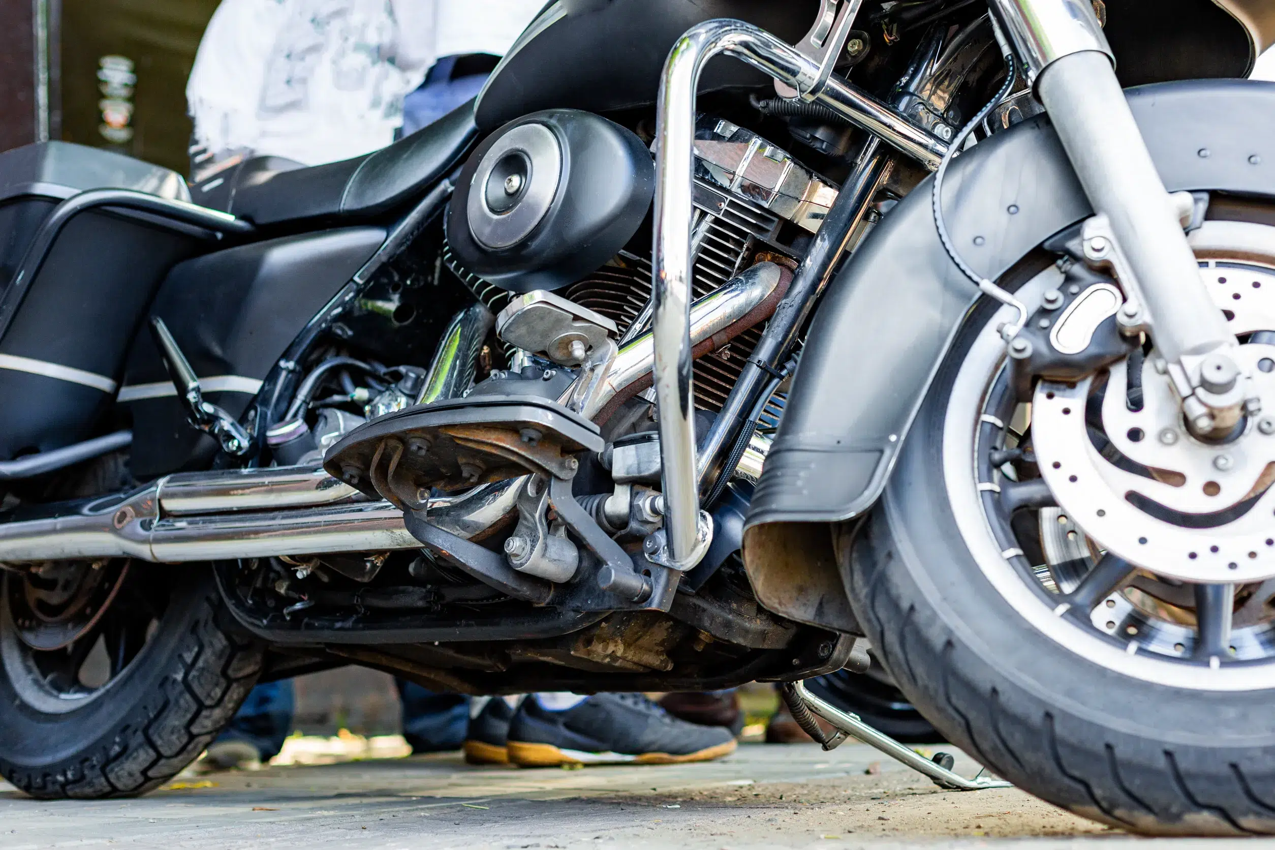 Close-up view of the engine and front wheel of a parked motorcycle, with part of a persons legs and feet visible—highlighting the importance of motorcycle safety equipment.
