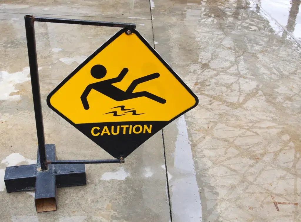 A yellow caution sign on wet concrete warns of a slipping hazard, displaying a stick figure slipping and the word “CAUTION.”.