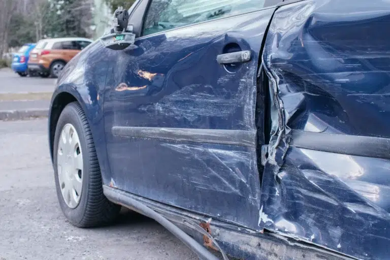 A blue car with significant damage and dents on the drivers side door, parked on a street with other vehicles in the background.
