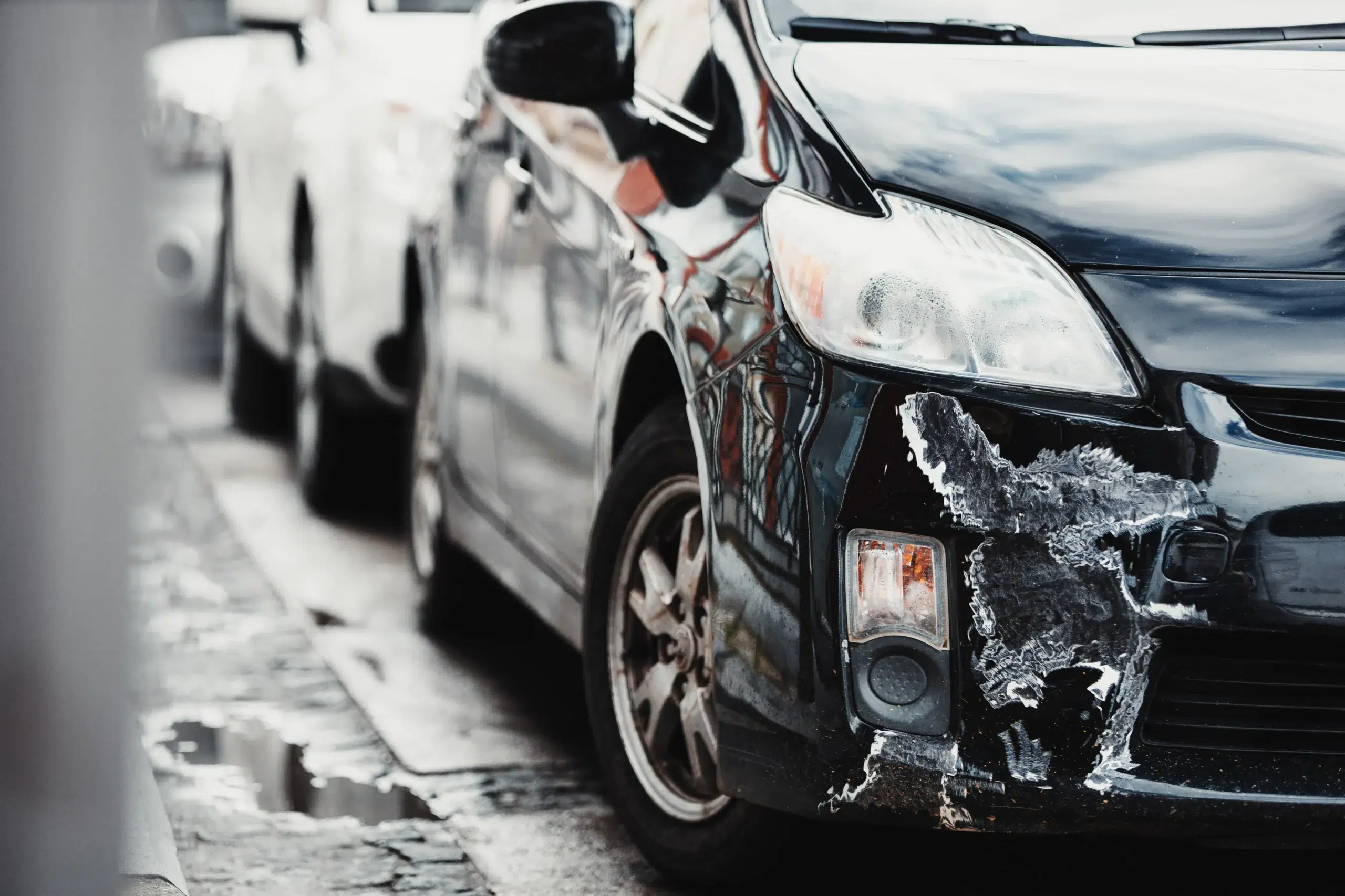 A black car with visible damage and scrapes on its front bumper is parked on a street next to a white vehicle.