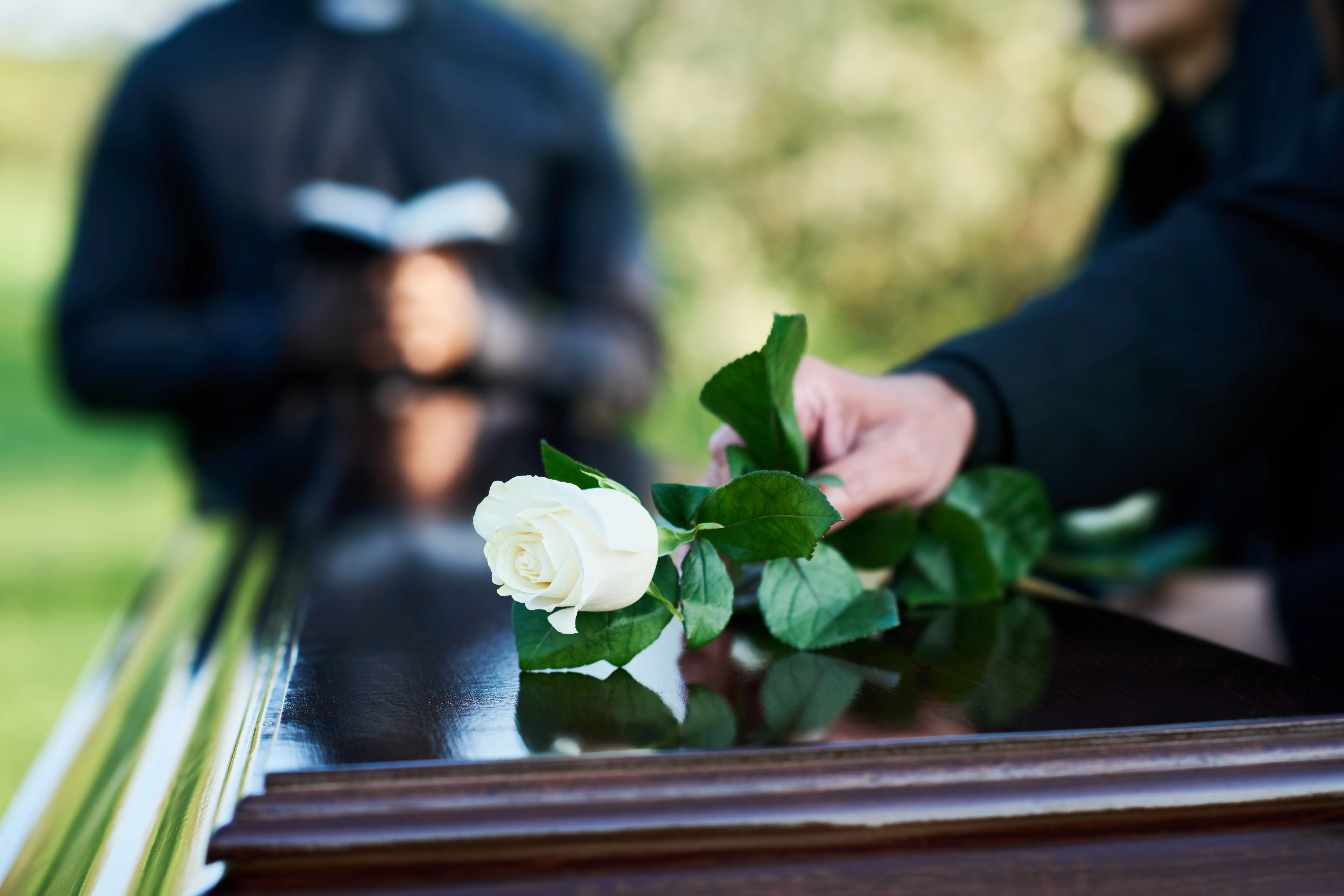 A person places a white rose on a polished wooden casket during a funeral ceremony, with another person holding an open book in the background.
