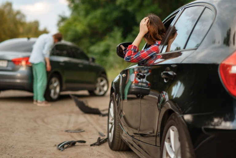 Two cars appear to have been in a collision on a rural road. A woman sits in one car holding her head, while a man inspects damage to the other vehicle. Debris is scattered on the ground.