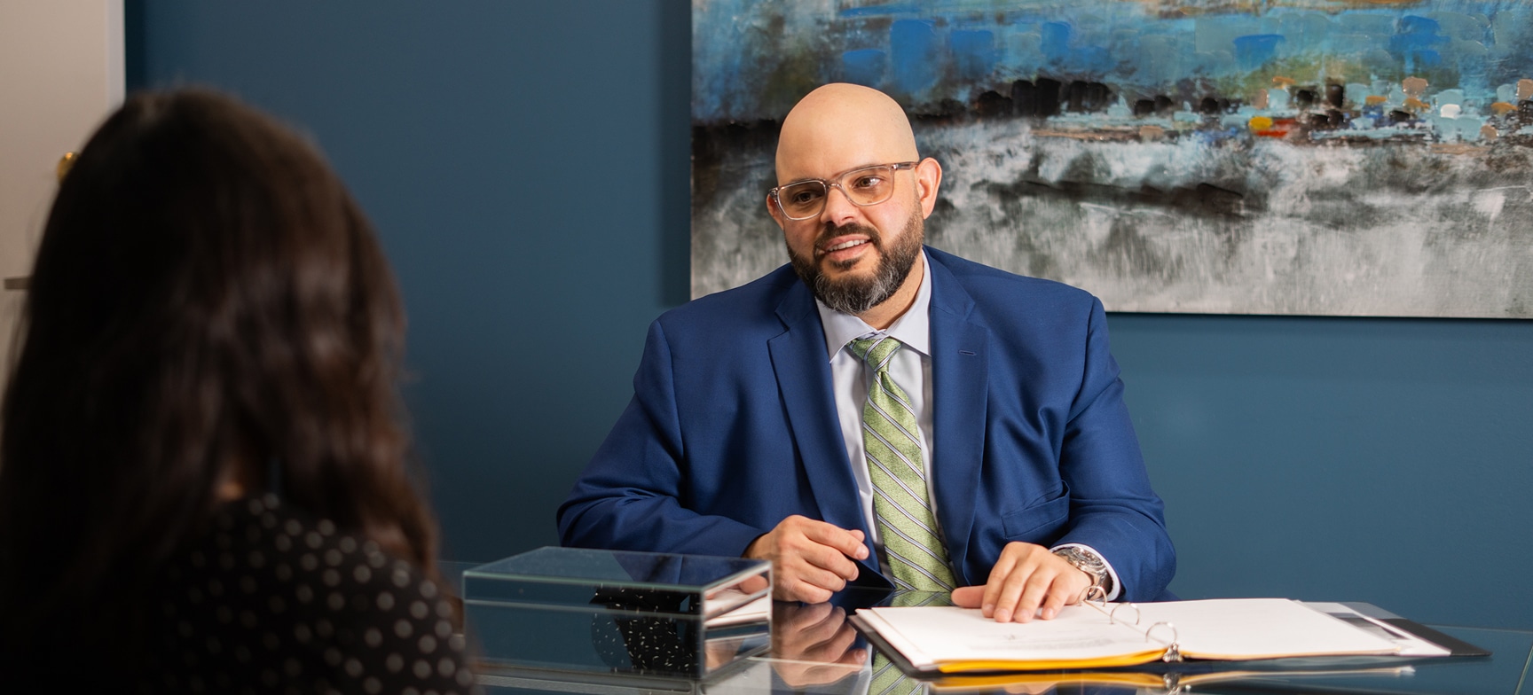 Attorney Roberto Vazquez in a blue suit and glasses sits at a desk talking to a woman. A painting is on the blue wall behind him.