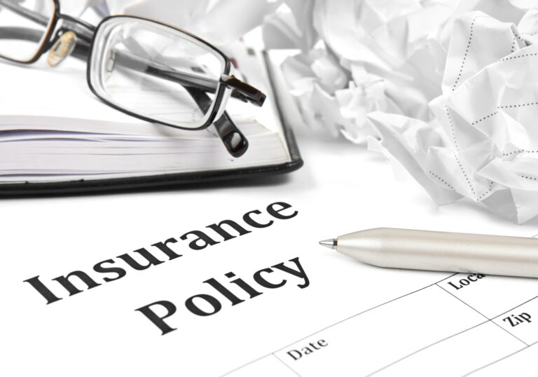 A pair of eyeglasses, a crumpled paper, and a pen rest on top of an insurance policy document and a notebook.