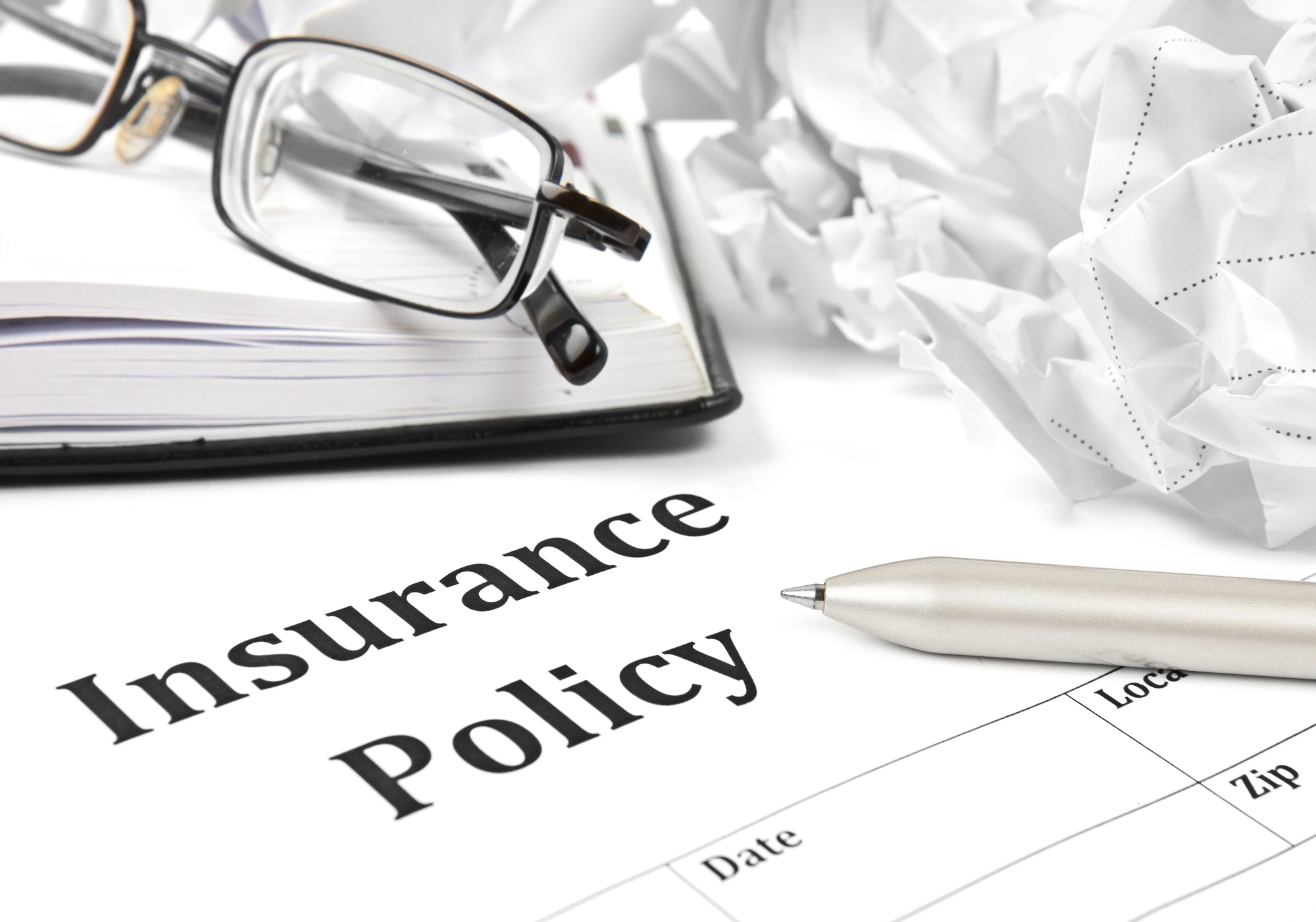 A pair of eyeglasses, a crumpled paper, and a pen rest on top of an insurance policy document and a notebook.