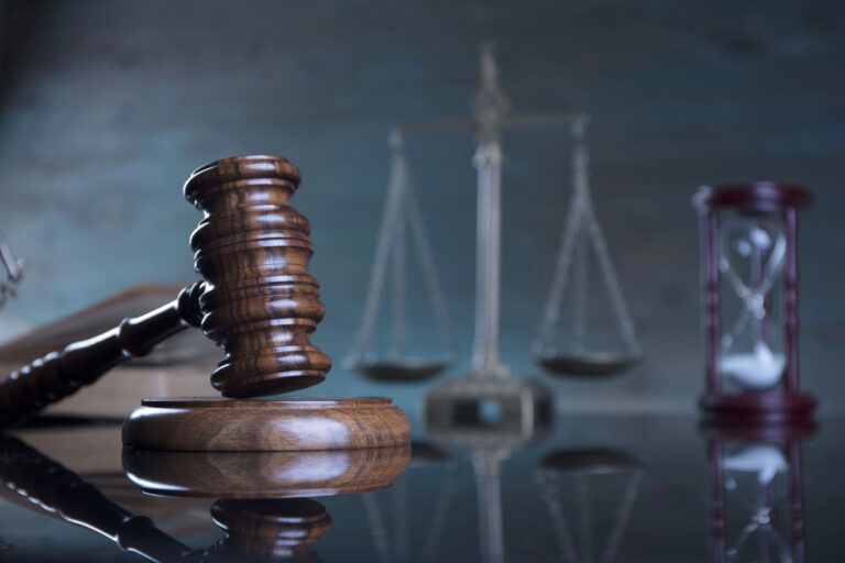 A wooden gavel rests on a sound block in front of blurred scales of justice and an hourglass, symbolizing law and justice.