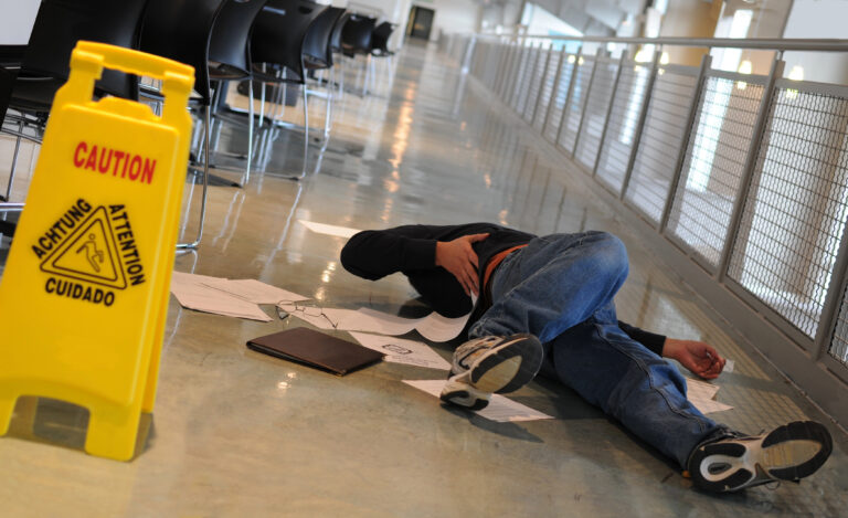 A person lies on the floor near scattered papers, clutching their side, next to a yellow caution sign indicating a slippery surface.