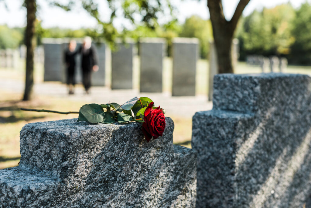 A single red rose rests on a gray gravestone in a cemetery, with two people standing in the blurred background among other gravestones.