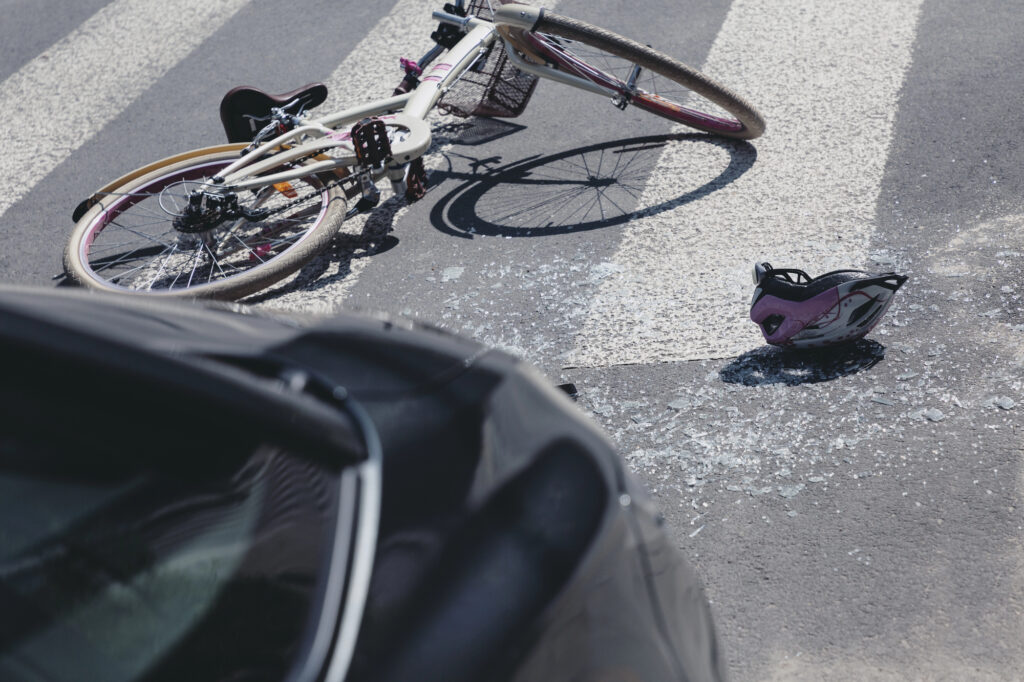 A bicycle and helmet lie on a crosswalk next to shattered glass, with the rear of a car visible in the foreground.