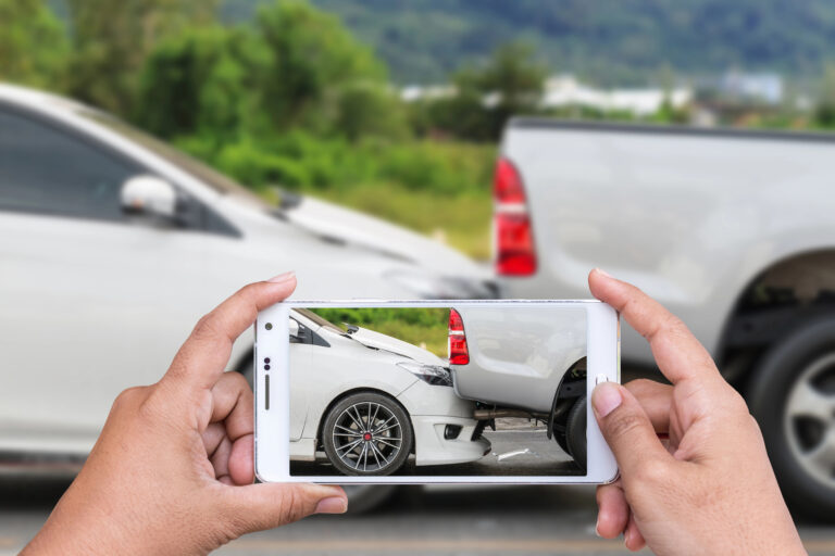A person takes a photo with a smartphone of a white car and a gray truck involved in a rear-end collision on a road.