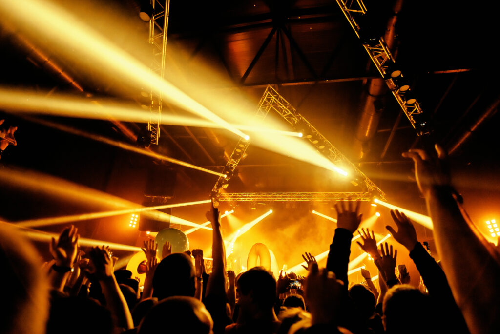 Crowd raising hands at a concert with bright yellow stage lights and geometric light structures overhead.