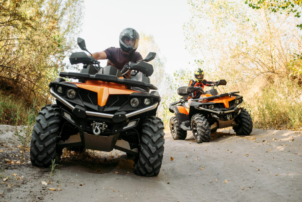 Two people wearing helmets are riding orange all-terrain vehicles (ATVs) on a sandy off-road trail surrounded by trees and shrubs.