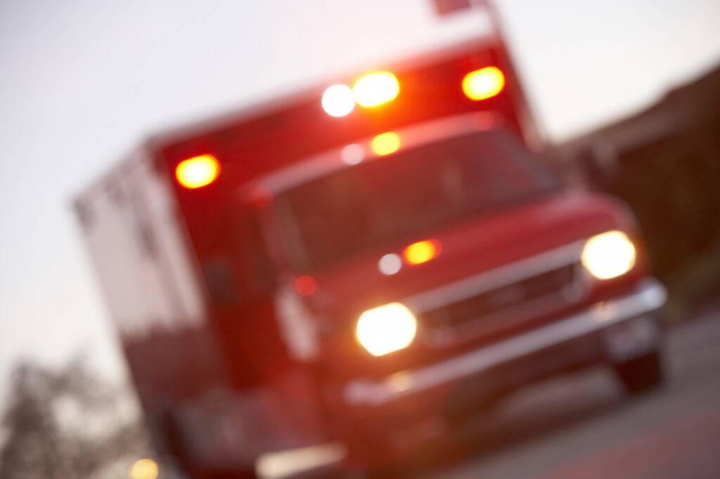 A red ambulance with flashing lights is shown out of focus, facing forward on a roadway.