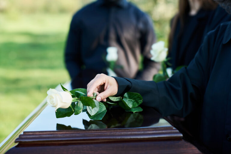 A person places a white rose on a wooden casket during an outdoor funeral service, with others holding roses in the background.