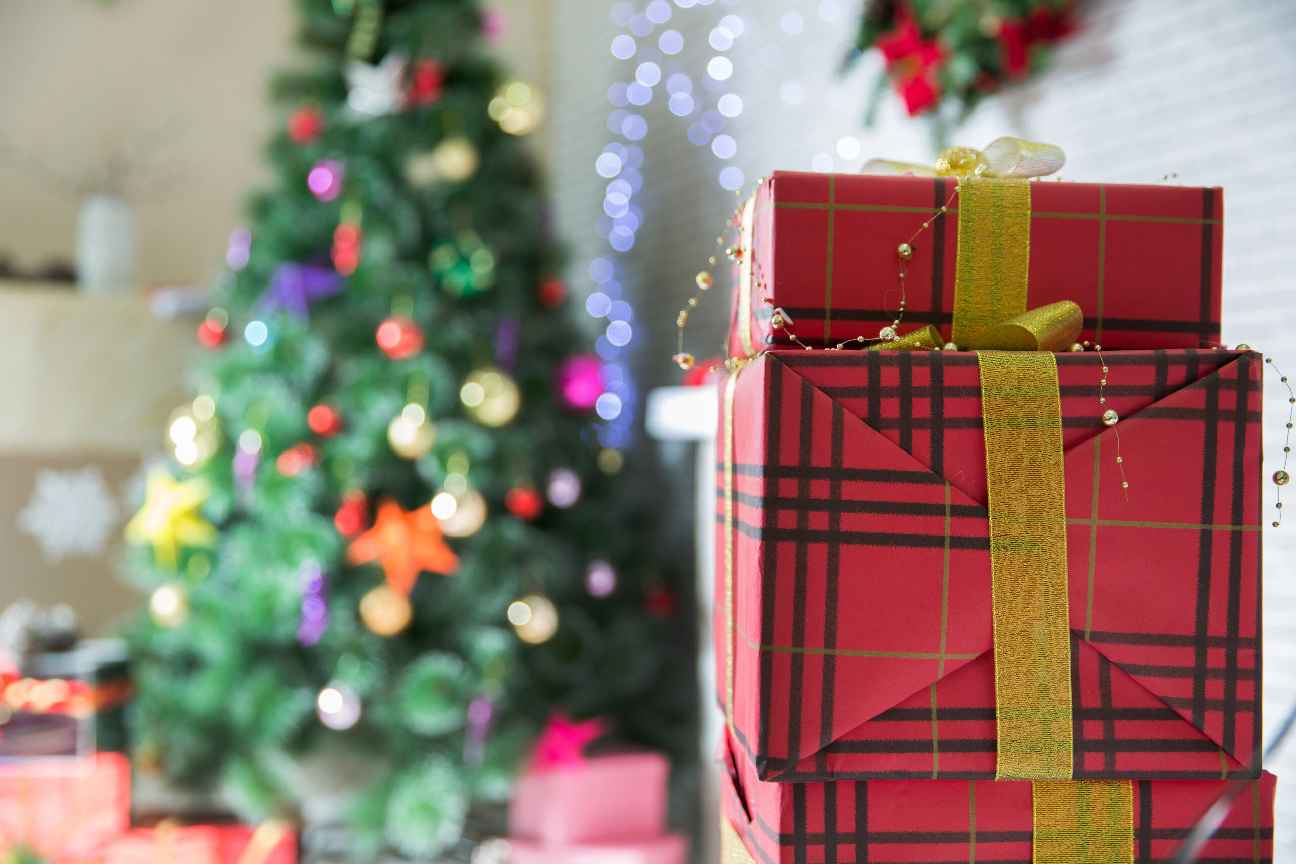Stack of red plaid gift boxes with gold ribbons in the foreground, with a decorated Christmas tree and holiday lights in the background.